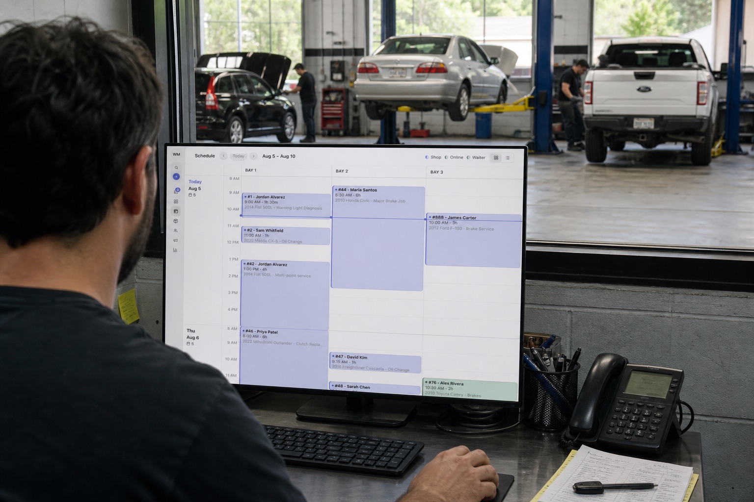 Service writer looking at a three-bay schedule on a monitor, with cars on lifts behind the glass